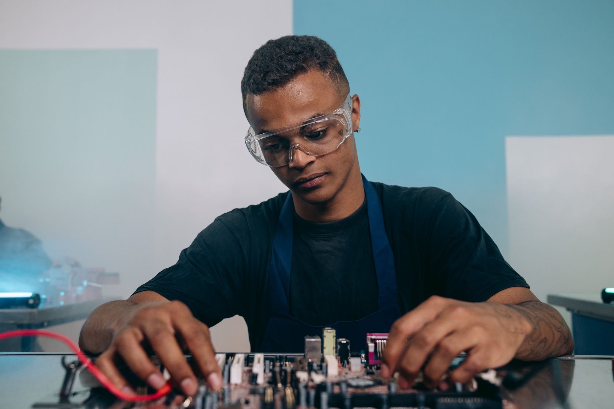 A young man wearing clear safety glasses and a dark apron works on an electronic circuit board at a bench.